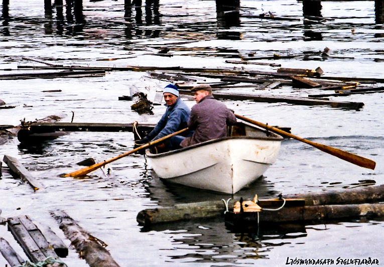 Unnið við að rífa ónýtar bryggjur. Bæjarstarfsmennirnir Jón á Hóli og Stebbi Andrésar róa í bryggjubrakinu og draga í land staura og annað drasl. Ljósmyndari: Steingrímur Kristinsson.